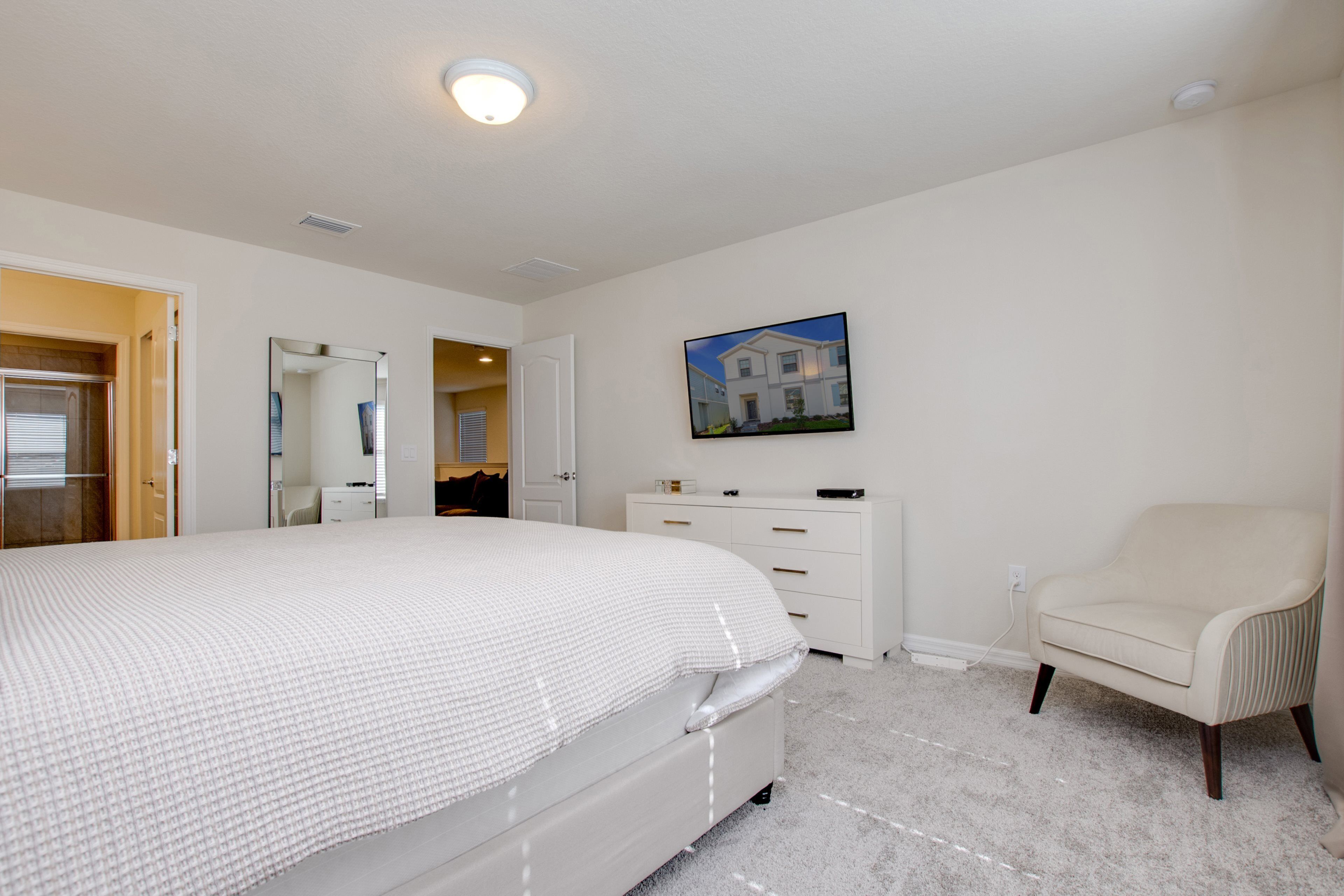 A bedroom with a large bed, a dresser, a chair, and a wall-mounted TV.