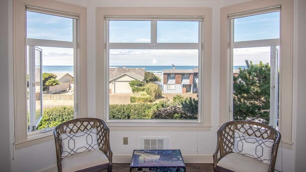 A room with three large windows offering a view of the beach and neighboring houses. There is a small table and two wicker chairs with cushions. - Ocean View Home! Hot tub! Dog Friendly! Minutes to the Beach! (Waldport)