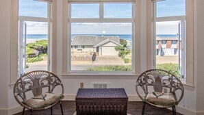 A room with three bay windows overlooking a beach and a small building. There are two wicker chairs with cushions. - Ocean View Home! Hot tub! Dog Friendly! Minutes to the Beach! (Waldport)