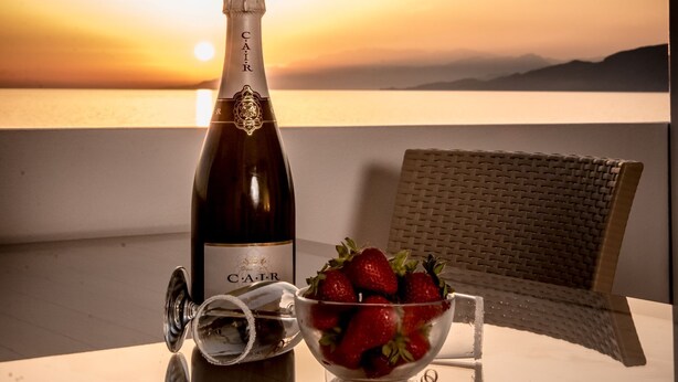 Une bouteille de champagne C.A.I.R., un saladier de fraises et une chaise en osier sur une table, le tout avec une vue sur le coucher de soleil.