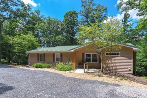 A single-story cabin with a green roof, surrounded by trees, a gravel driveway, and a small porch area. - Quiet Lakefront w/ Deck, Dock & Fire Pit (Swanton)