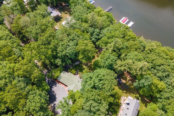 An aerial view of a residential area with dense greenery, a small house with a green roof, and a dock extending into a lake. - Quiet Lakefront w/ Deck, Dock & Fire Pit (Swanton)