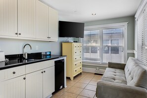 Cocina moderna con armarios blancos, encimera negra y lavavajillas integrado. Hay un televisor montado sobre la encimera y una ventana con persianas a la derecha.