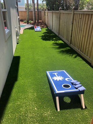 A backyard with artificial grass, a shuffleboard table, and a wooden fence. - 2 Rooftop Decks | Downtown IOP | Pool | Elevator | 1 Blk From Beach | Pets (Isle of Palms)