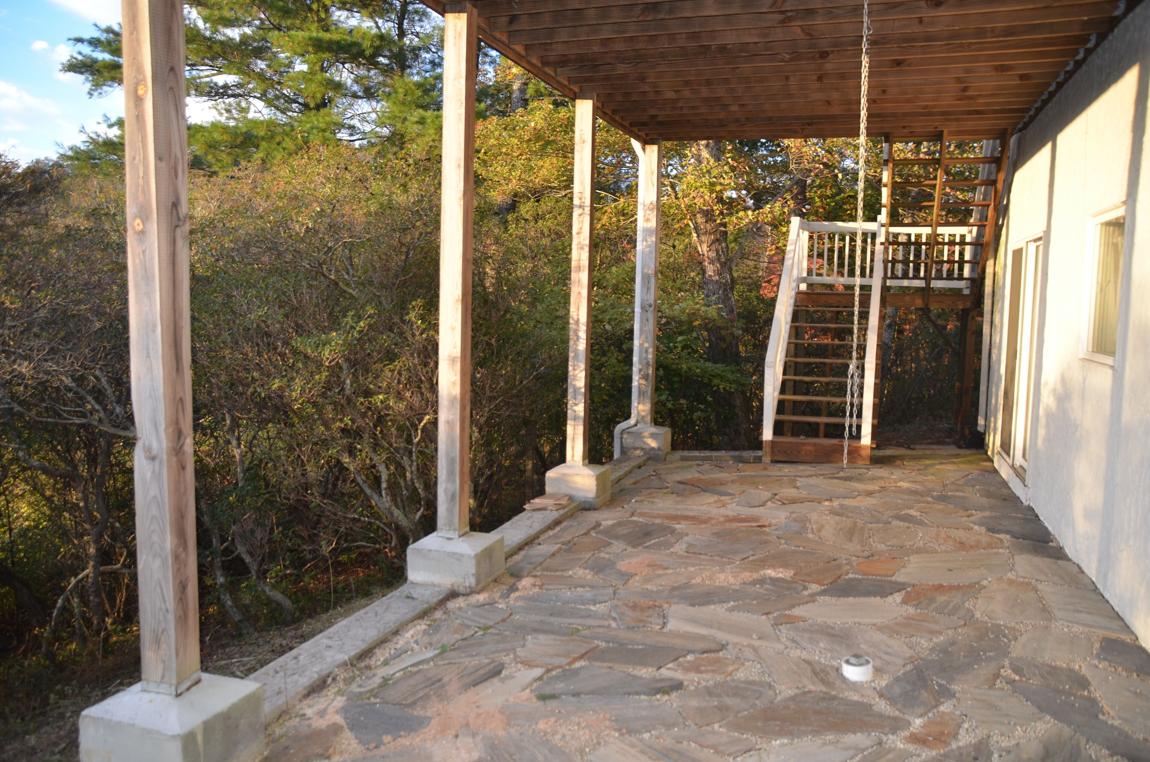 A stone-paved porch with wooden columns, a wooden staircase, and a view of a forested area.