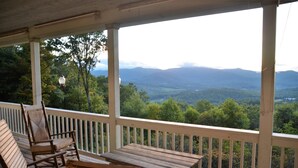 Una terraza de madera con una mesa y sillas, con vista a un valle boscoso y montañas a lo lejos.