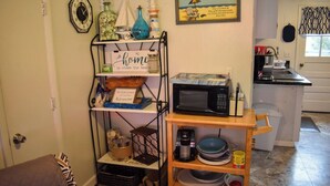 Una cocina con microondas, cafetera y lavamanos. Hay una estantería con diversos utensilios de cocina y un letrero que dice "home is where the beach is".