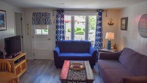 Una sala de estar acogedora con un sofá azul, una mesa de centro de madera con una cita motivacional, un televisor sobre un mueble de madera y una puerta que da al exterior.