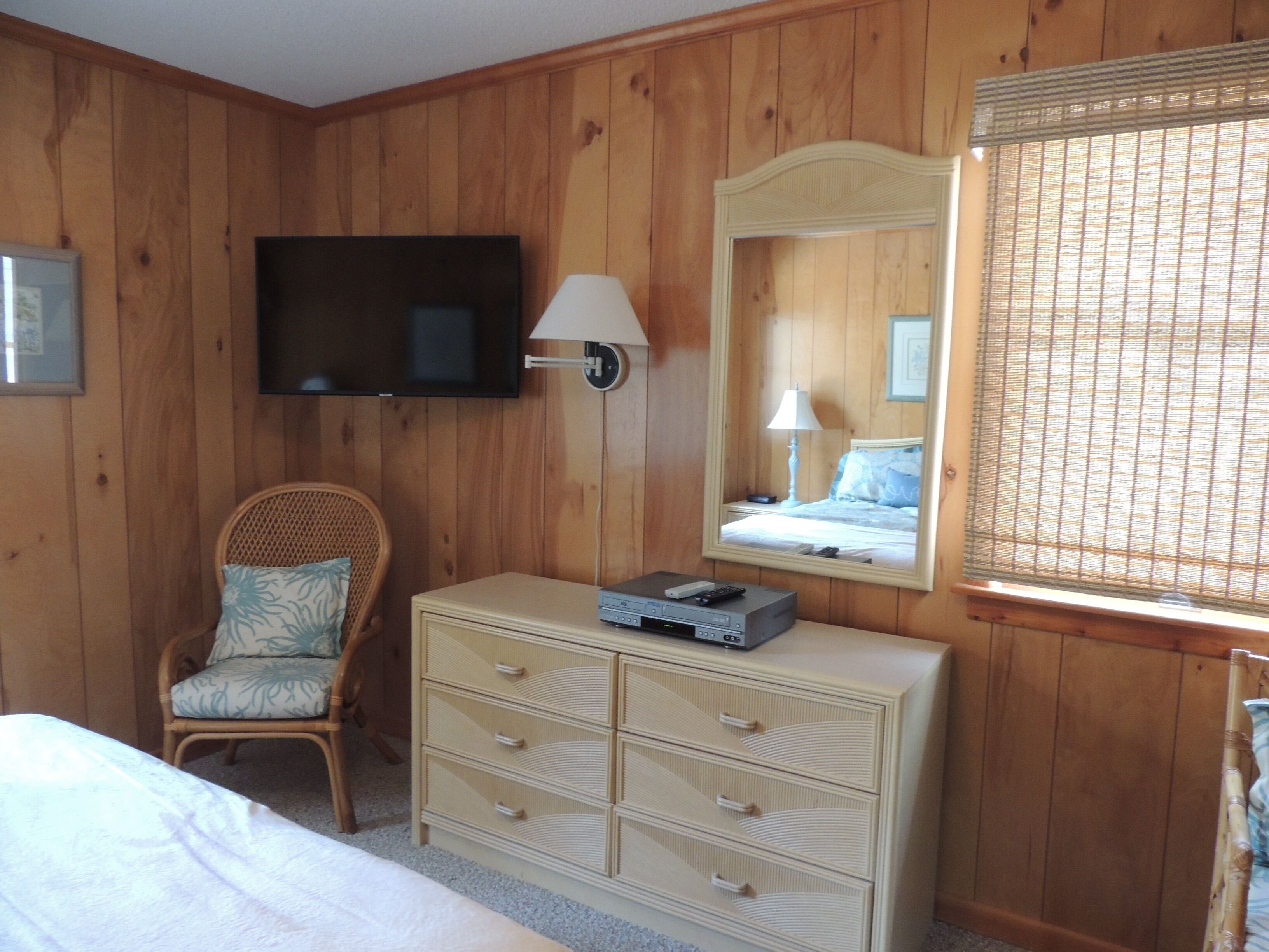 A wooden-paneled room with a bed, a wicker chair, a dresser with drawers, a television mounted on the wall, and a window with blinds.