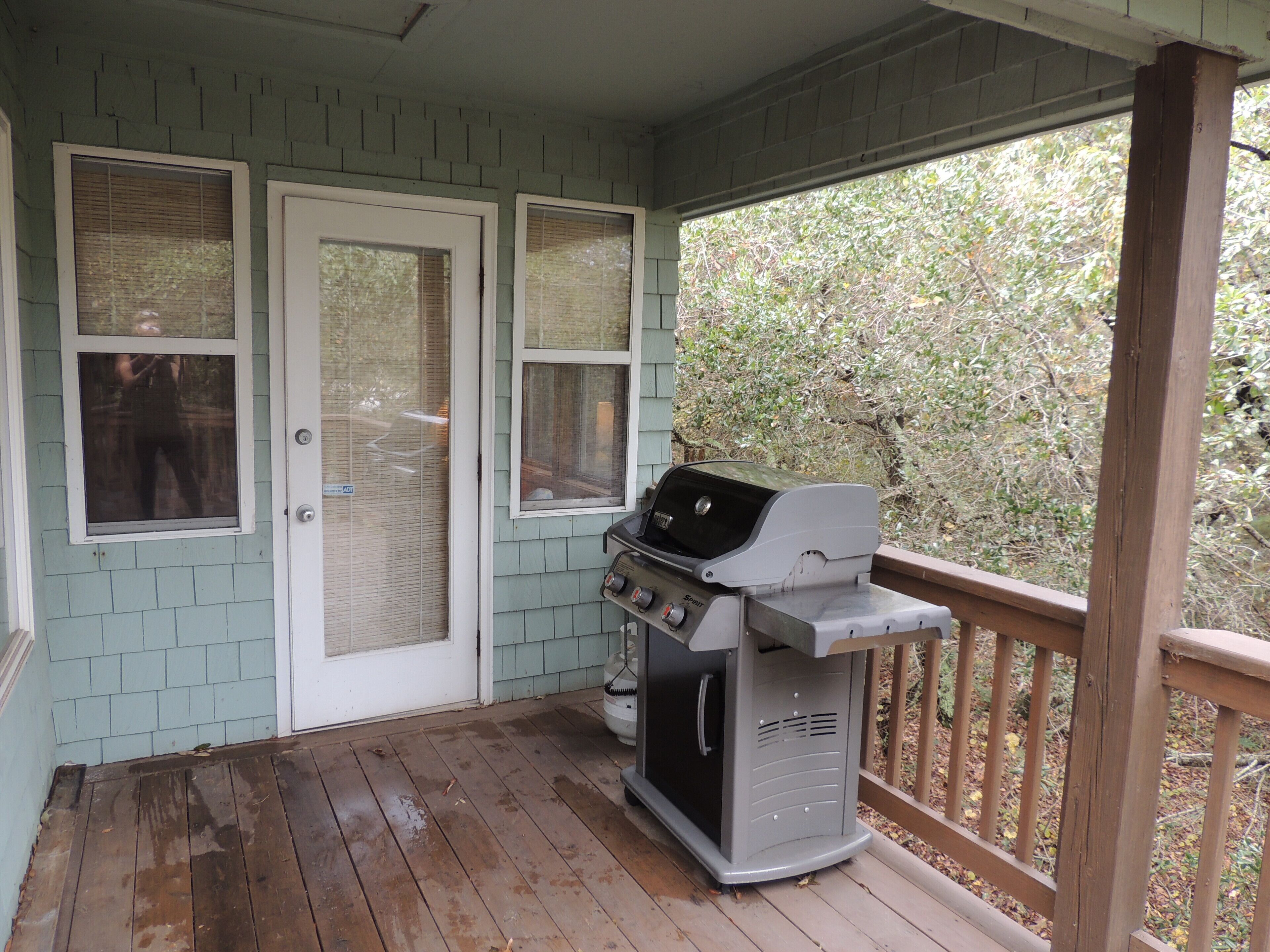 A covered porch with a grill, wooden deck, and a view of trees.