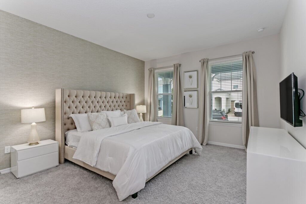 A bedroom with a tufted headboard, a bed with white linens, a nightstand with a lamp, a TV on a stand, and a window with curtains.