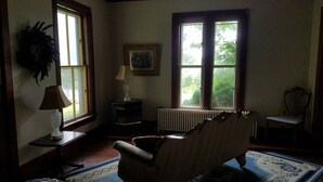 Una habitación con radiador, una silla, una mesa con lámpara y dos ventanas que dan a zonas verdes.