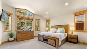 A bedroom with a large bed, wooden headboard, a dresser, and a nightstand. There are multiple windows providing natural light and views of the outdoors. - Deck, Bikes, Newer Build in Upper Kings Beach (Kings Beach)