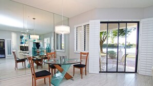 Uma área de jantar com mesa e cadeiras de vidro, um candelabro e vista para uma área externa através de uma porta de vidro.