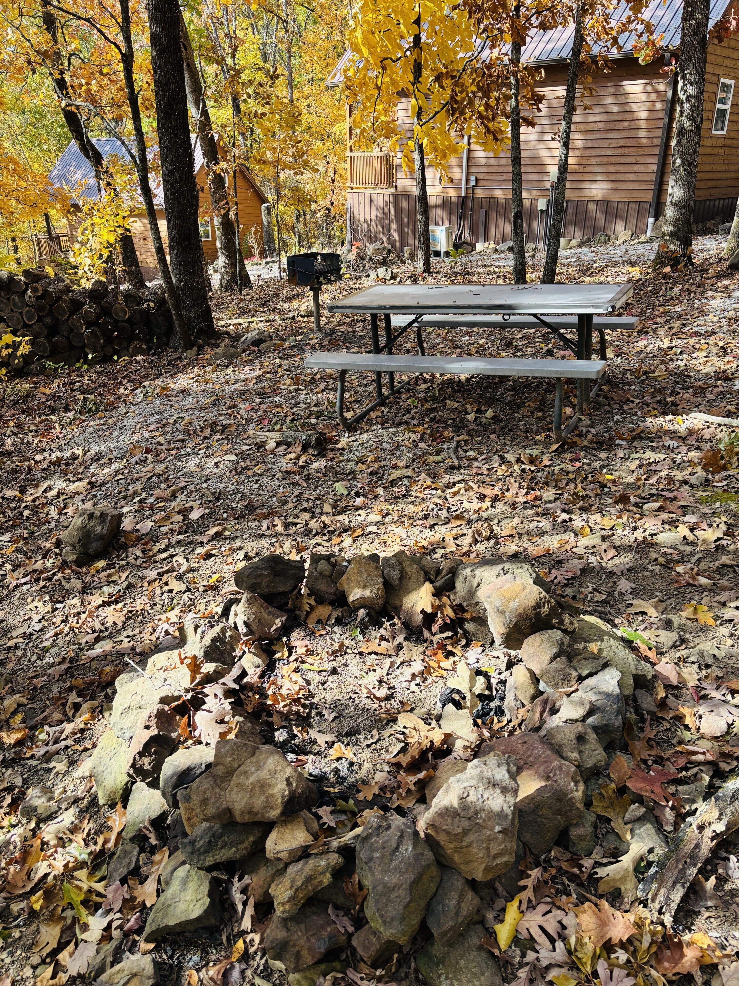 Un hogar de piedra rodeado de hojas otoñales, una mesa de picnic y una cabaña de madera al fondo.