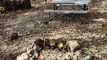 Un hogar de piedra rodeado de hojas otoñales, una mesa de picnic y una cabaña de madera al fondo.