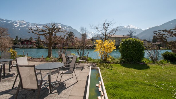 Une terrasse avec des sièges extérieurs et un petit élément décoratif aquatique, offrant une vue sur un lac avec des montagnes en arrière-plan.