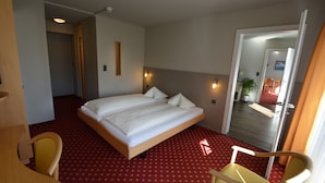 Une chambre d’hôtel avec un grand lit, un bureau et une chaise. Une porte donne sur une autre pièce, et la fenêtre offre une vue sur un couloir agrémenté d’une plante.