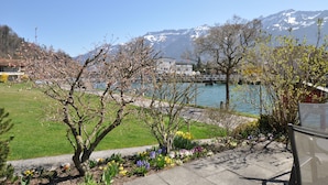 Une vue sur le lac, avec des montagnes en arrière-plan, une zone herbeuse parsemée de plantes en fleurs et un bâtiment doté d’un toit rouge.