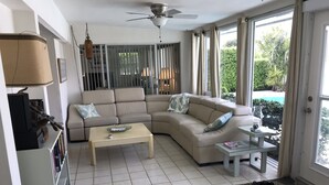 A living room with a beige sectional sofa, a wooden coffee table, a small side table, and a door leading to a balcony with a view of a pool. - Bright Spacious 3bed/2bath Intracoastal Home (Deerfield Beach)