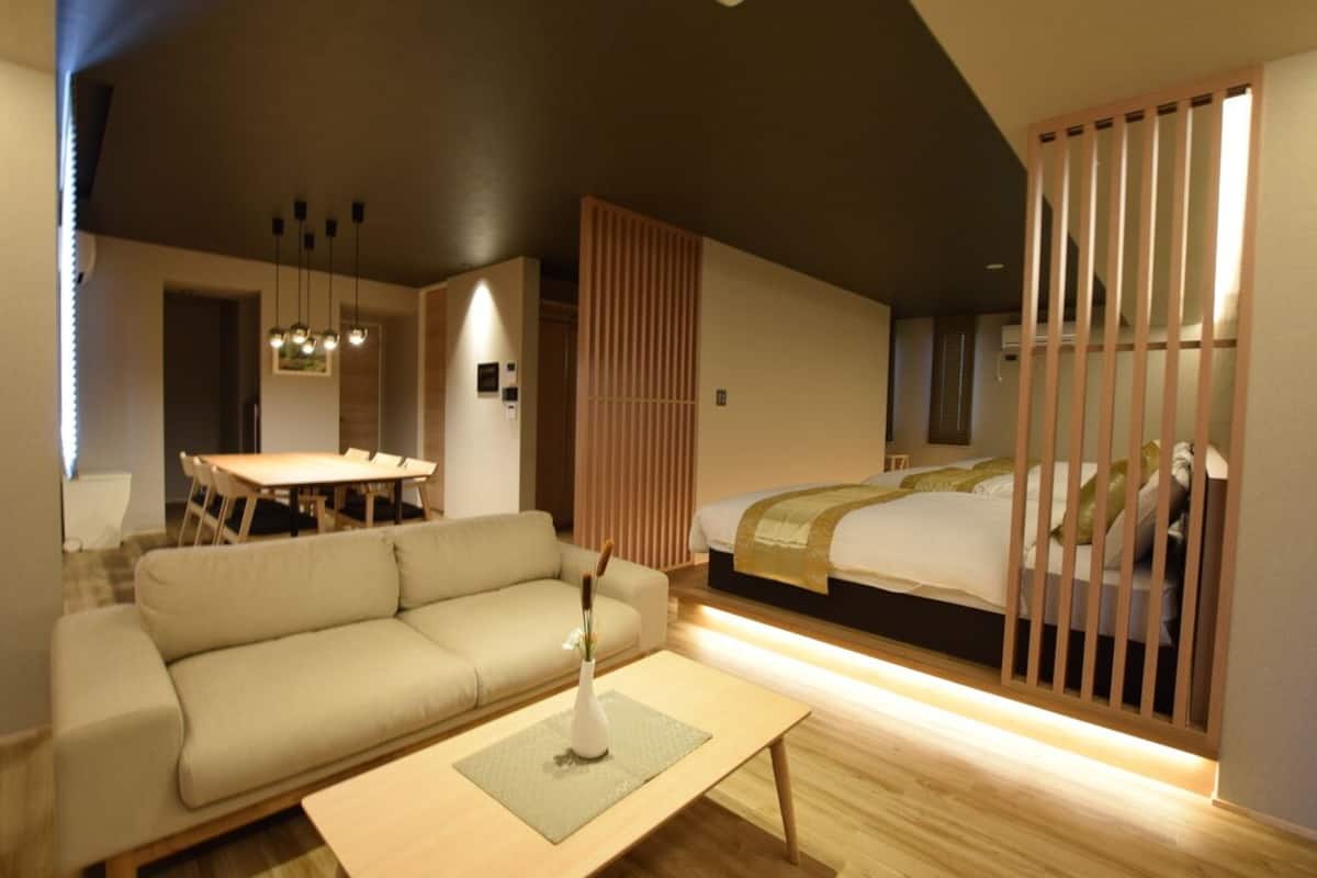 A modern hotel room with a sofa, a dining table, a bed with a wooden headboard, and a partition with vertical slats.
