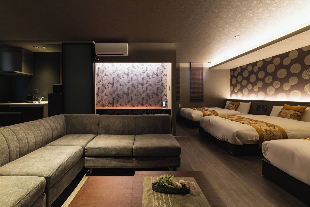 Ein modernes Hotelzimmer mit einer Eckcouch, einem Bett mit gemustertem Kopfteil, einem kleinen Tisch mit einer Pflanze und Blick auf die Stadt durch ein Fenster.