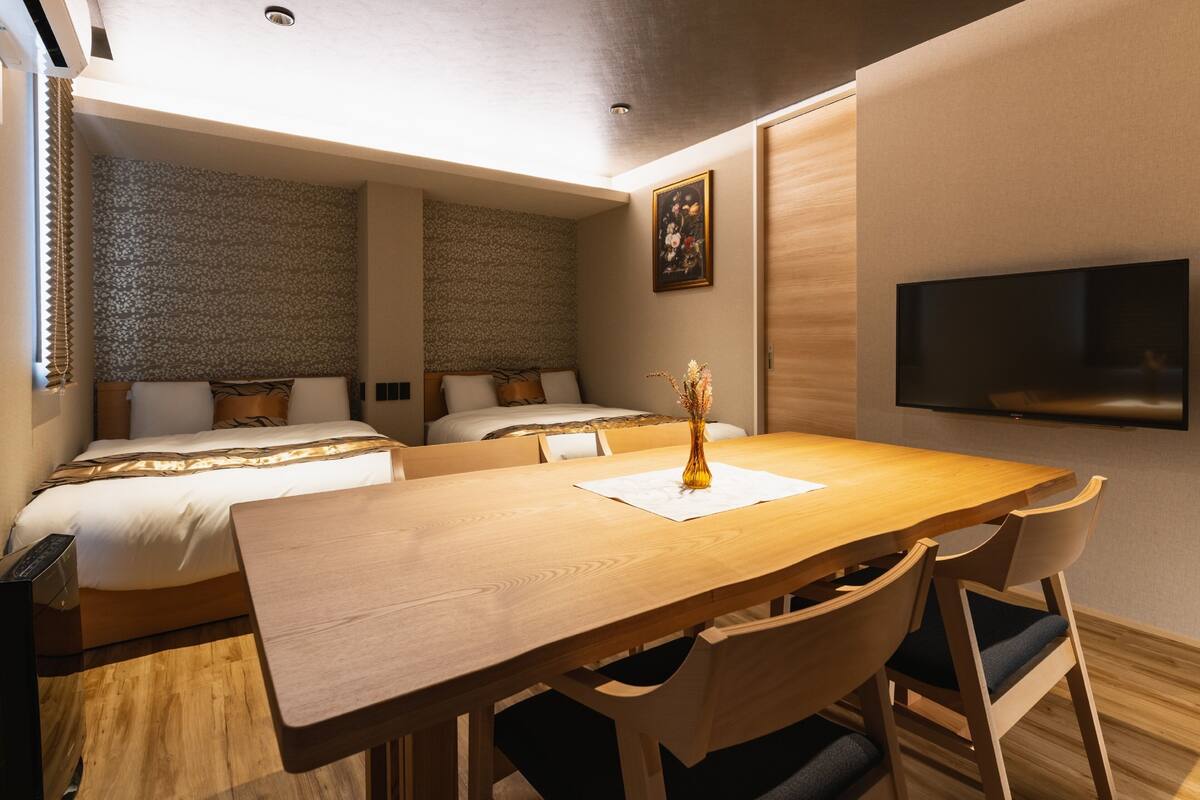 Ein Hotelzimmer mit zwei Betten, einem Holztisch und einem an der Wand befestigten Flachbildfernseher.