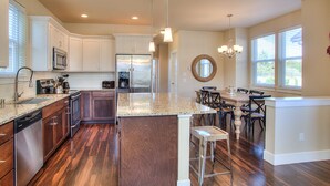 A modern kitchen with wooden cabinets, a granite countertop, stainless steel appliances, and a dining area with a round table and chairs. - Windswept Cottage - Oyhut Bay Seaside Village (Ocean Shores)