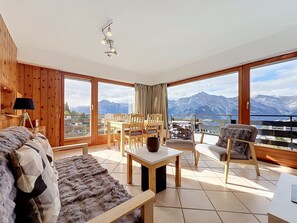 Ein geräumiger Raum mit großem Fenster und Blick auf die Berge, einer gemütlichen Sitzecke mit Sofa und Sesseln sowie einem Esstisch.
