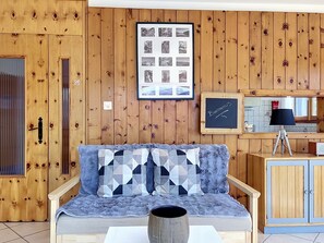 Ein Zimmer mit Holzwänden, einer Couch, einem Couchtisch, einer Collage gerahmter Fotos und einer Tafel mit dem Schriftzug 'Bienvenue'.