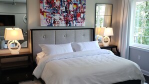 Un dormitorio moderno con una cama grande, dos mesitas de noche con lámparas, un ventilador de techo, un cuadro abstracto de grandes dimensiones y una ventana con cortinas.