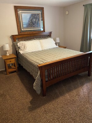 A bedroom with a wooden bed, two bedside tables with lamps, a framed picture on the wall, and a carpeted floor.