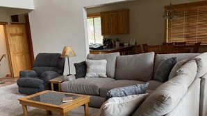 A living room with a grey sofa, a wooden coffee table, and a small wooden side table. There is a wooden door and a window with blinds in the background.