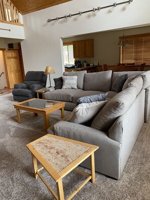 A living room with a grey sofa, a wooden coffee table, and a small wooden side table. There is a wooden door and a window with blinds in the background.