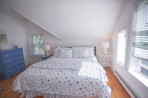 A bedroom with a sloped ceiling, a bed with a star-patterned bedspread, a blue dresser, a bedside table with a lamp, and a window with blinds.