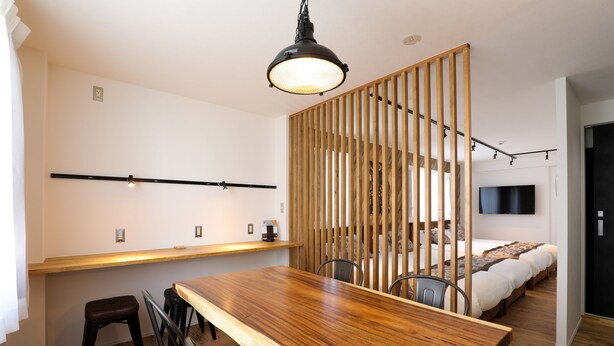 Una habitación moderna con una mesa de comedor de madera, sillas, un separador de madera, una cama con televisor y piso de madera.