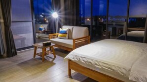 Habitación de hotel con una cama grande, un sofá con cojines, una mesita de madera y un ventanal con vistas a la ciudad.