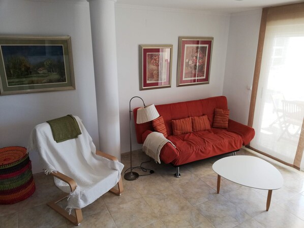 Un salon avec un canapé rouge, un fauteuil blanc, une petite table blanche et un lampadaire. Deux tableaux encadrés ornent le mur, et une fenêtre offre une vue sur des meubles d’extérieur.