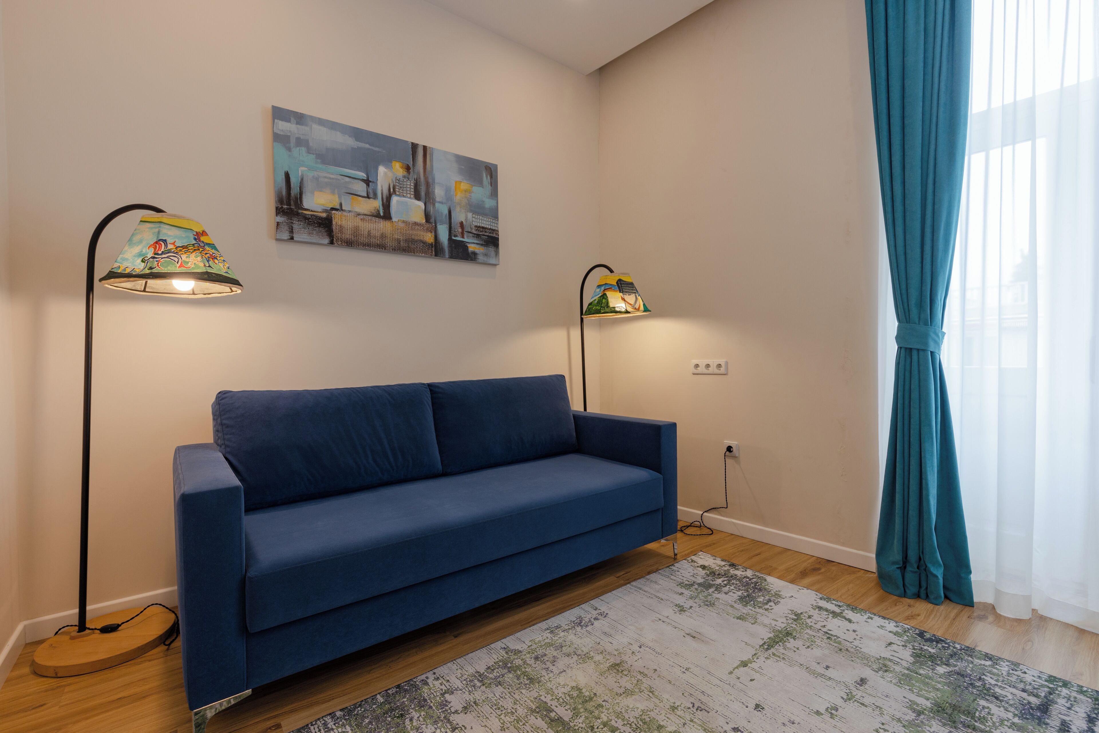 A modern living room with a blue sofa, two floor lamps, a patterned rug, and a cityscape painting on the wall.