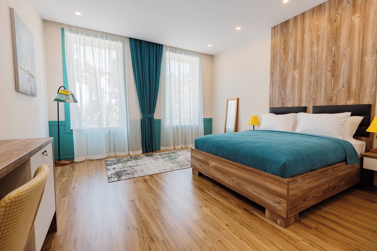Une chambre à coucher avec un lit en bois, un bureau, une chaise et une grande fenêtre avec des rideaux.