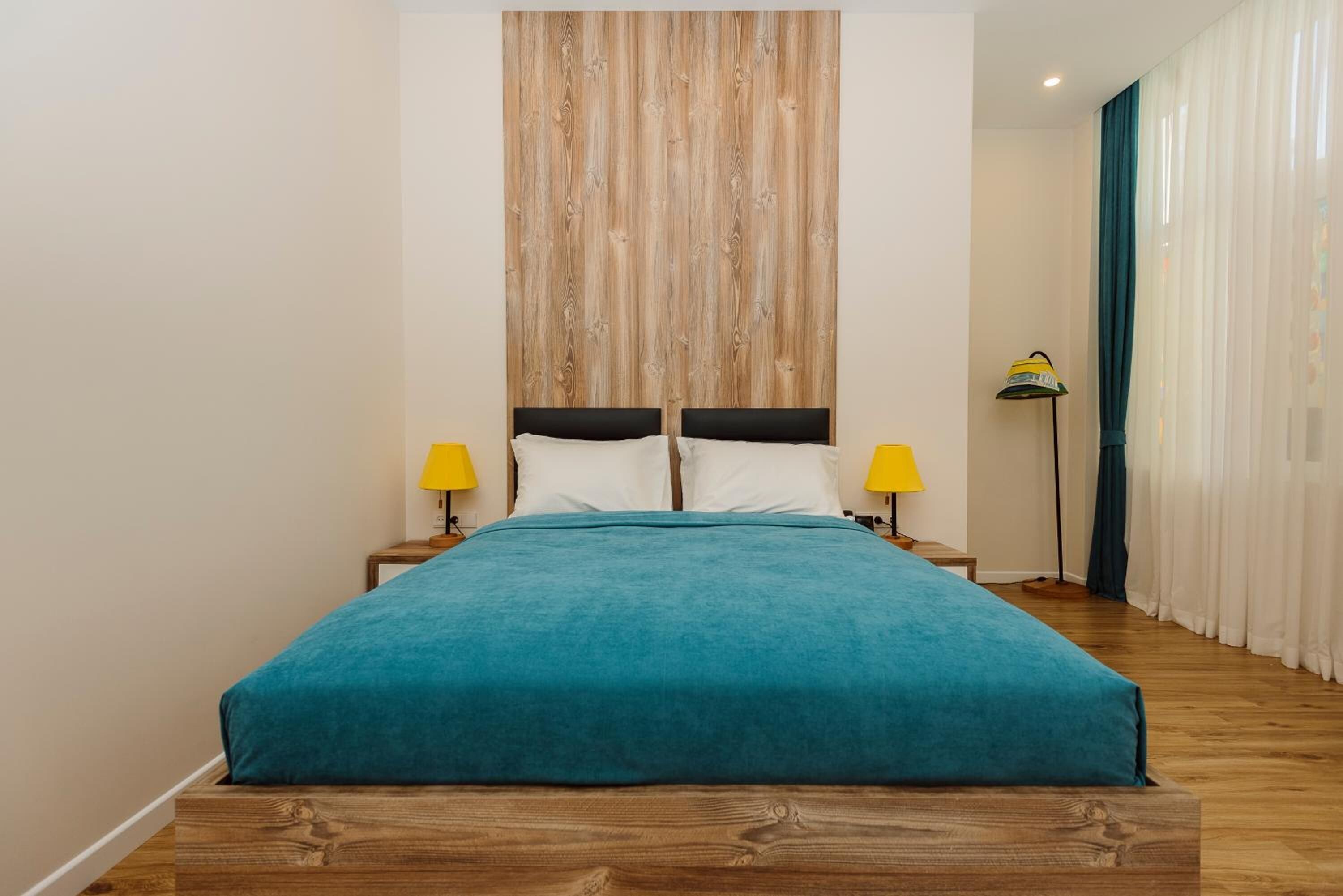 A bed with a teal bedspread, wooden headboard, and two bedside tables with yellow lamps.