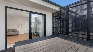 A modern outdoor deck with a wooden floor, a black mesh privacy screen, and a glass door leading to an interior space with a sofa and a desk.