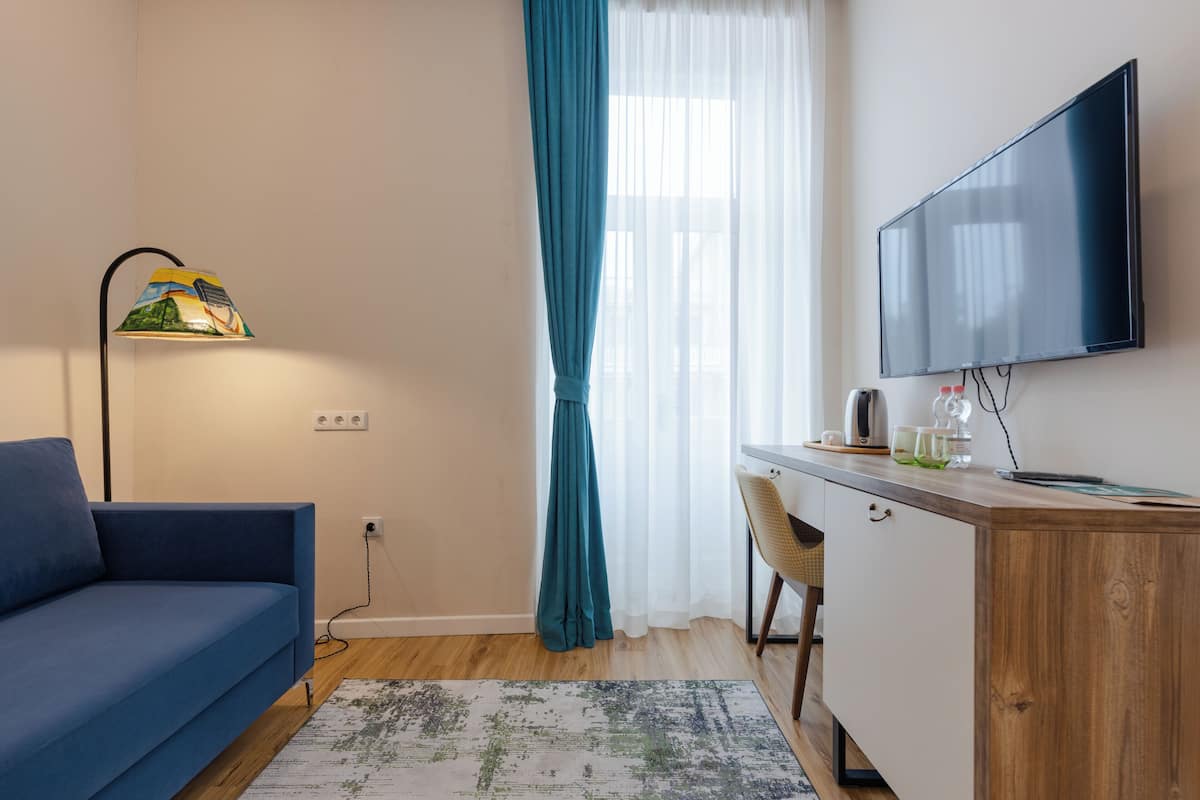 Un salon moderne avec un canapé bleu, un bureau doté d’un téléviseur à écran plat, une lampe avec un abat-jour coloré et une grande fenêtre ornée de rideaux légers.