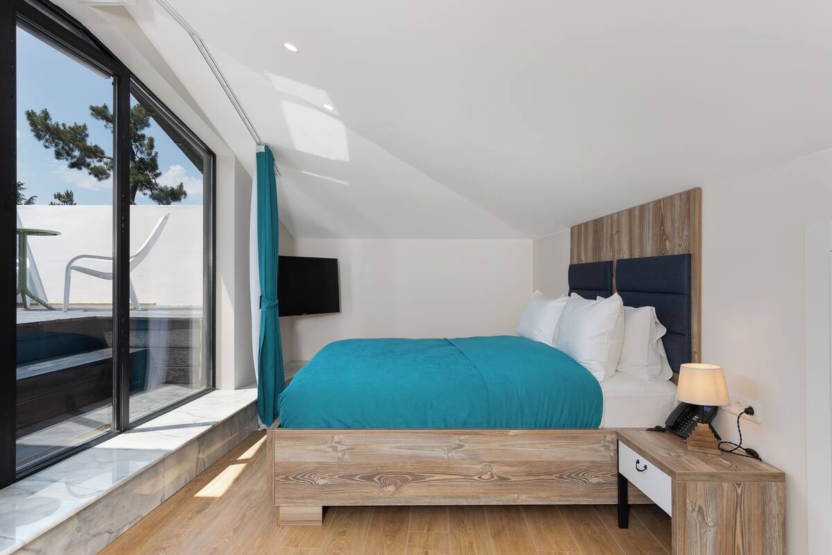 Une chambre moderne avec un grand lit, une table de chevet en bois, une télévision et une vue sur l’extérieur grâce à une grande fenêtre.