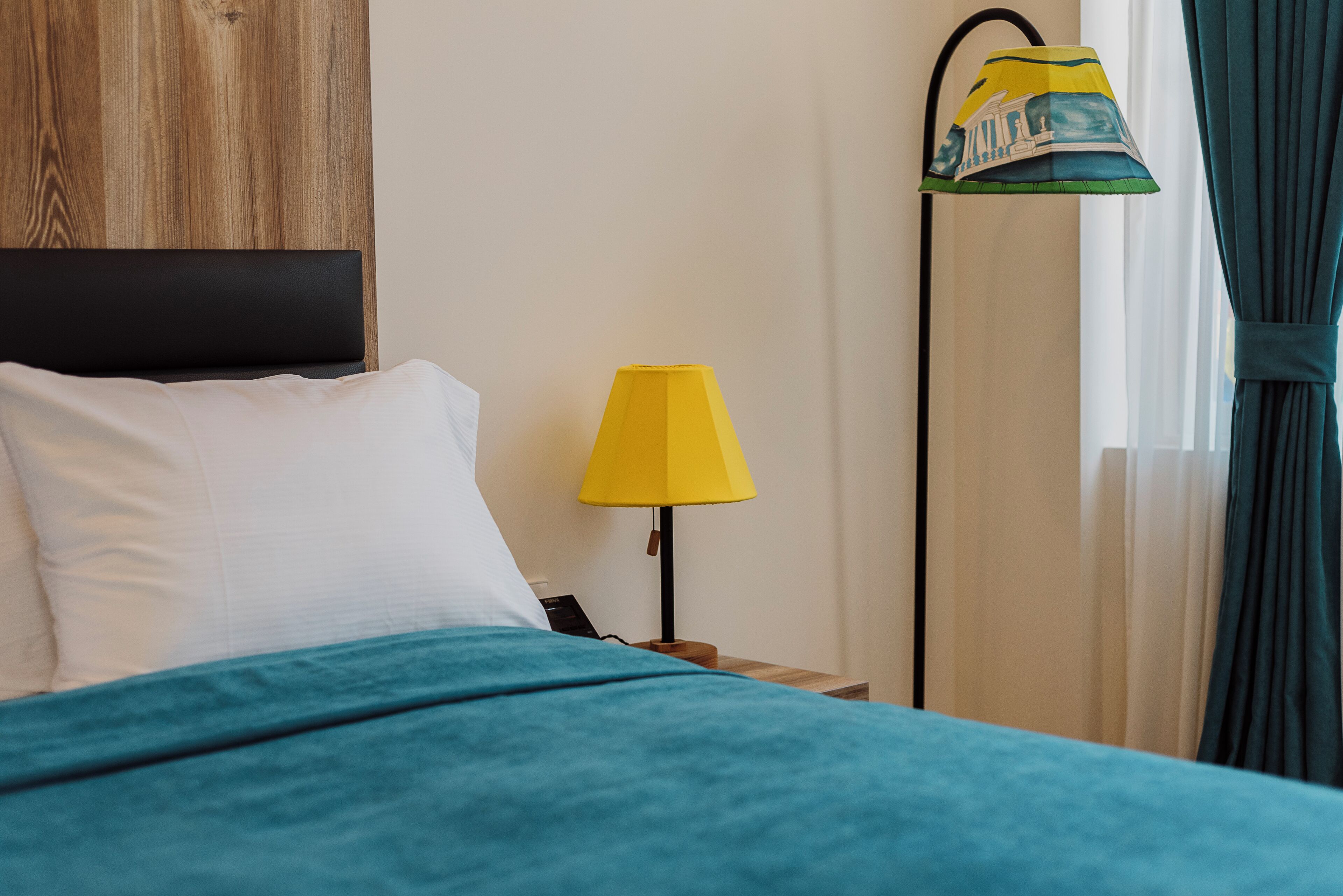 A neatly made bed with a teal bedspread and white pillows, a yellow lamp on a wooden nightstand, and a window with sheer curtains.
