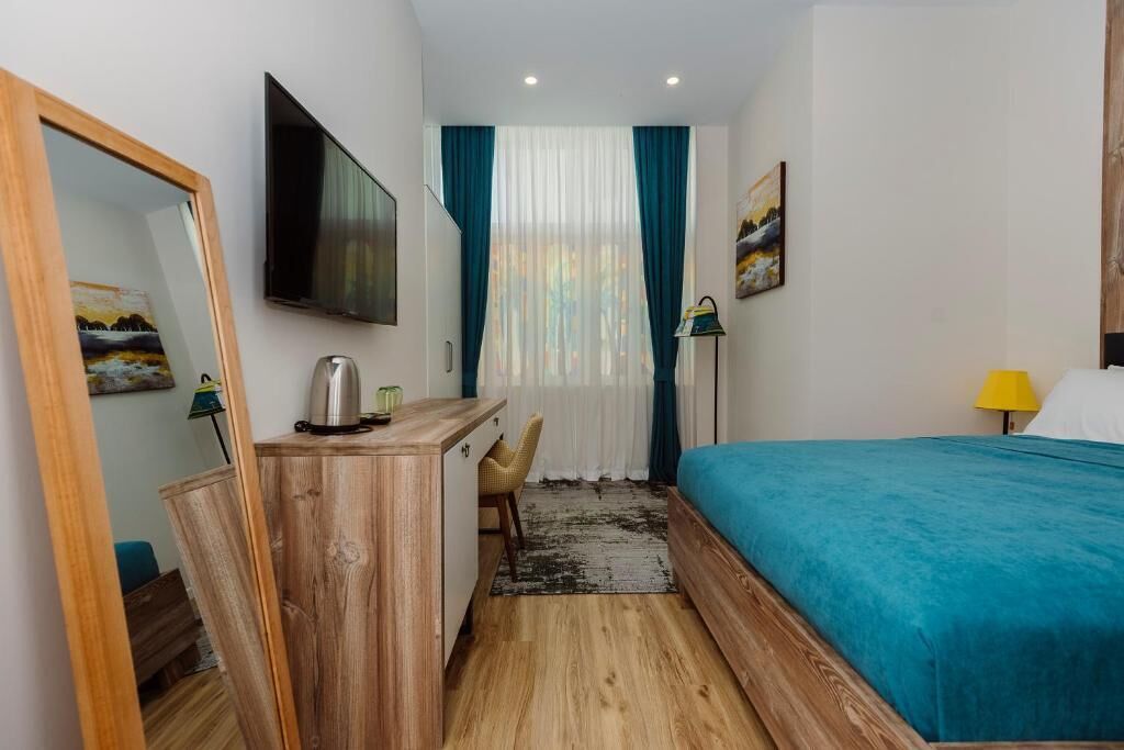 A hotel room with a bed, a desk with a kettle, a chair, a TV, and a window with curtains.
