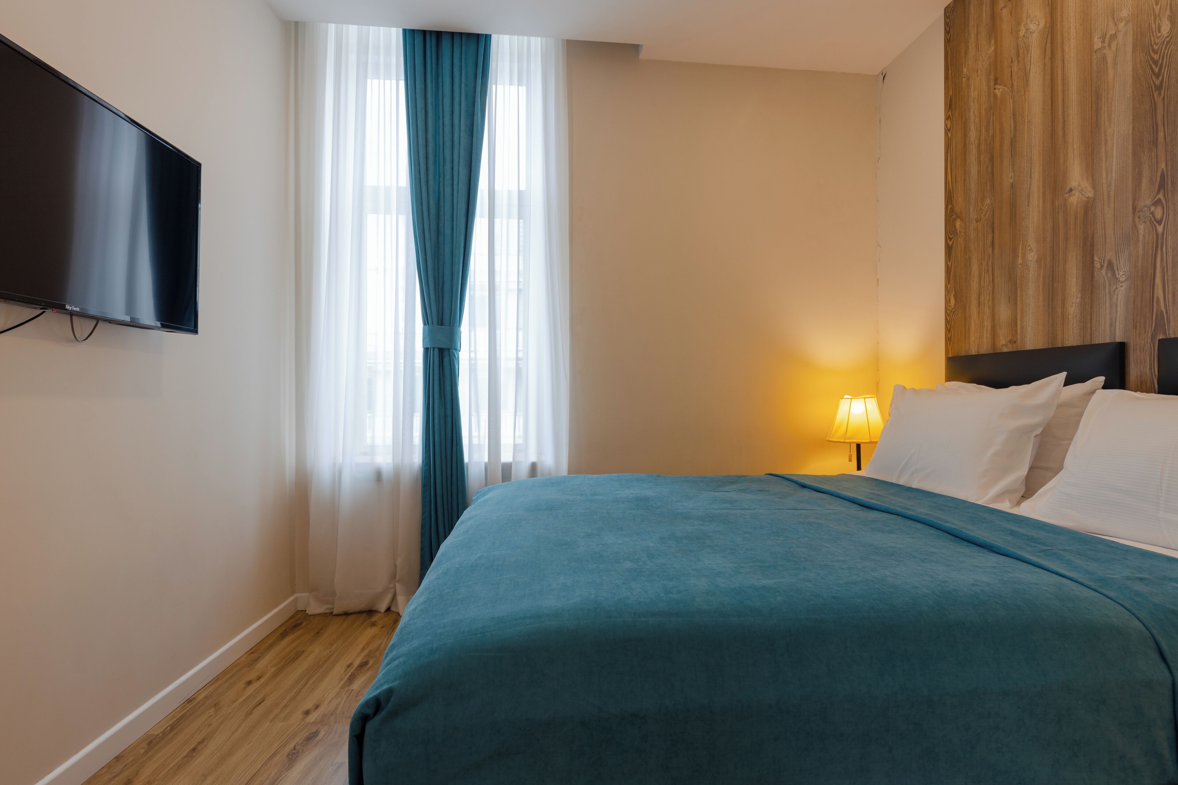 Ein Hotelzimmer mit Bett, Wandfernseher, Fenster mit Vorhängen und einer Holzpaneelausstattung an der Wand.