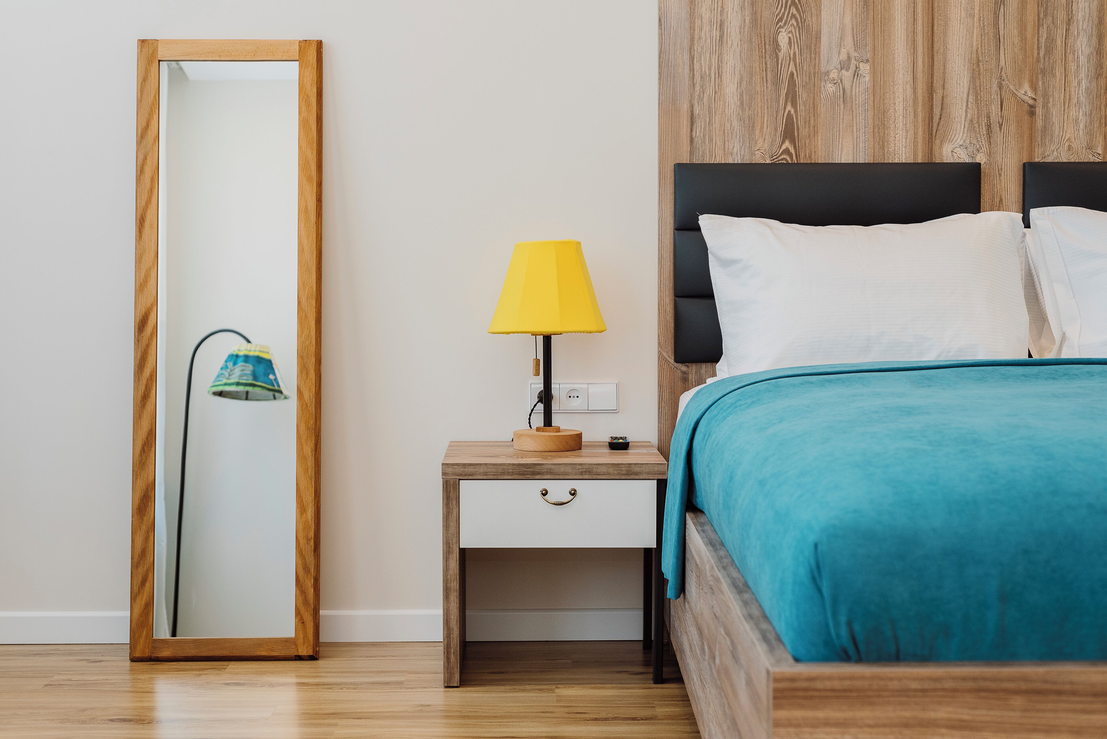 Un dormitorio con una mesita de noche de madera, una lámpara amarilla, una cama con una manta color turquesa y un cabecero de madera.