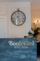 A reception desk with a clock above it and a vase of flowers to the right. The text "Boulevard Hotel Batumi" is visible on the desk.