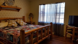 A bedroom with a wooden bed, a quilt, a television on a wooden stand, a window with sheer curtains, and a wall-mounted shelf with a decorative item. - Backyard splendor!!! (Story)